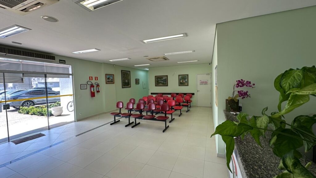 A imagem mostra a sala de espera de um consultório odontológico. As paredes são pintadas em um tom claro de verde-água. No chão, há um piso de ladrilhos brancos com algumas linhas azuis decorativas. À esquerda, há uma grande porta de vidro que dá para a rua, onde se pode ver carros estacionados e uma bicicleta. Acima da porta de vidro, há uma placa com a palavra "SAÍDA" em letras brancas. Na parede à esquerda, perto da entrada, há dois extintores de incêndio vermelhos e uma placa. Há também algumas pinturas nas paredes, com molduras escuras. A sala de espera está equipada com várias cadeiras vermelhas dispostas em fileiras. As cadeiras parecem ser feitas de metal e plástico. No canto direito da imagem, há um balcão com uma decoração de plantas, incluindo uma orquídea roxa em um vaso e uma planta verde grande com folhas largas.