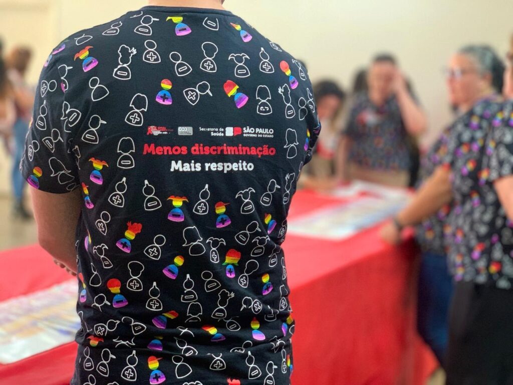 A imagem mostra a parte de trás de uma pessoa usando uma camiseta preta com um padrão repetido de figuras humanas estilizadas em branco e algumas figuras coloridas com as cores do arco-íris. Essas figuras coloridas parecem representar diversidade e inclusão, possivelmente relacionadas à comunidade LGBTQ+. No centro da camiseta, há uma mensagem impressa em branco e vermelho que diz: "Menos discriminação, Mais respeito". Acima dessa mensagem, aparecem os logotipos e nomes da "Secretaria de Saúde" e do "Governo do Estado de São Paulo", indicando que a camiseta faz parte de uma campanha ou ação oficial do governo. O cenário ao fundo está desfocado, mostrando outras pessoas e uma mesa vermelha, sugerindo que a foto foi tirada em um evento ou reunião pública. A iluminação é natural e o foco principal está na camiseta e na mensagem que ela transmite.