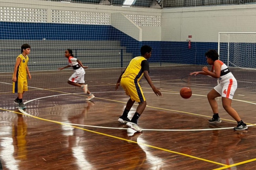 A imagem mostra um jogo de basquete indoor com quatro jovens jogadores em ação. No centro da imagem, dois jogadores estão se enfrentando. Um jogador vestindo um uniforme amarelo e preto com listras azuis está com a bola, driblando-a com a mão direita. Ele está olhando para frente, possivelmente para um companheiro de equipe ou para a cesta. Do outro lado, um jogador com um uniforme branco e laranja, com uma faixa preta no peito e o número 4 visível na perna, está em posição defensiva, com as mãos levantadas para tentar roubar a bola. À esquerda, um outro jogador com o mesmo uniforme amarelo e preto do jogador que está com a bola está parado, observando o jogo. Mais atrás, um quarto jogador, vestindo um uniforme branco e laranja semelhante ao do defensor central, está correndo, possivelmente em direção à cesta ou para se posicionar em quadra. O piso da quadra é de madeira brilhante, refletindo a luz. As linhas da quadra, em amarelo e branco, delimitam a área de jogo. Ao fundo, vê-se a arquibancada com cadeiras azuis e brancas, e redes de proteção. Há também uma cesta de basquete visível no canto superior direito da imagem. A iluminação parece ser artificial, típica de ginásios esportivos
