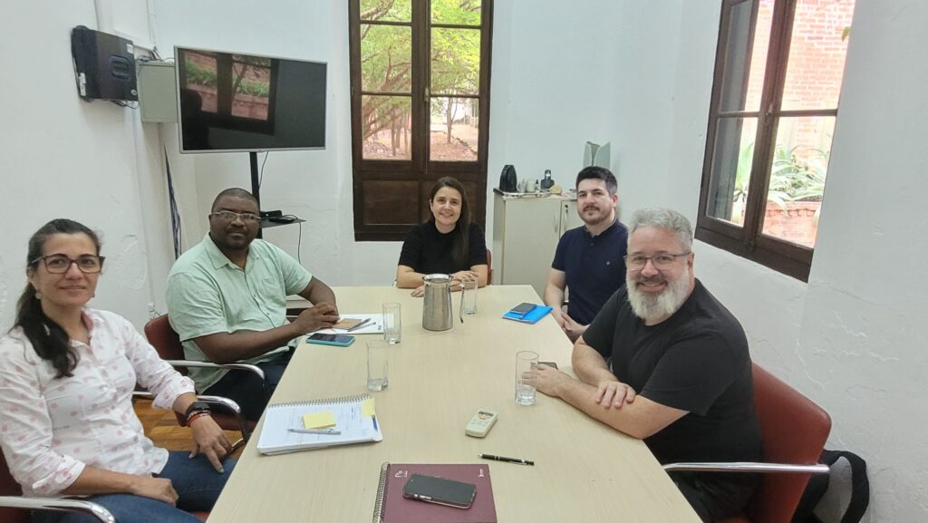 A imagem mostra um grupo de cinco pessoas sentadas ao redor de uma mesa em uma sala. Há uma televisão montada na parede à esquerda e uma grande janela com venezianas de madeira no centro da sala. As pessoas parecem estar em uma reunião ou discussão, com papéis e telefones celulares espalhados pela mesa. À esquerda, uma mulher com óculos e cabelo escuro está sorrindo para a câmera. Ao lado dela, um homem com óculos e camisa verde clara está sentado. No centro, uma mulher com cabelo escuro e uma blusa preta está sorrindo. À sua direita, um homem com uma camisa azul escura está olhando para a câmera. Finalmente, à direita, um homem com barba grisalha e óculos, vestindo uma camisa preta, está sorrindo para a câmera. A atmosfera geral da imagem é profissional e colaborativa.
