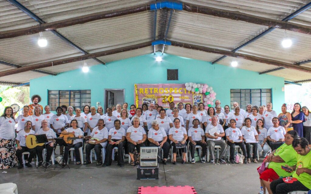 A imagem mostra um grande grupo de pessoas, a maioria idosas, reunidas em um salão com teto de estrutura metálica e telhas aparentes. Ao fundo, uma parede azul clara exibe um banner com a inscrição "RETROSPECTIVA 2005 - 20 ANOS", decorado com balões coloridos. A maioria das pessoas veste camisetas brancas com um logo vermelho e amarelo, possivelmente de uma instituição ou evento. Algumas pessoas estão sentadas em cadeiras, enquanto outras estão em pé, formando várias fileiras. Há instrumentos musicais visíveis, como um violão e um acordeão, sugerindo que o evento pode ter incluído apresentações musicais. No lado esquerdo da imagem, um homem com cabelo crespo e barba, usando uma camisa colorida, parece ser o fotógrafo ou um organizador. No lado direito, duas mulheres mais jovens, uma delas usando um vestido azul escuro, parecem estar interagindo com o grupo. A atmosfera geral é de celebração e confraternização.