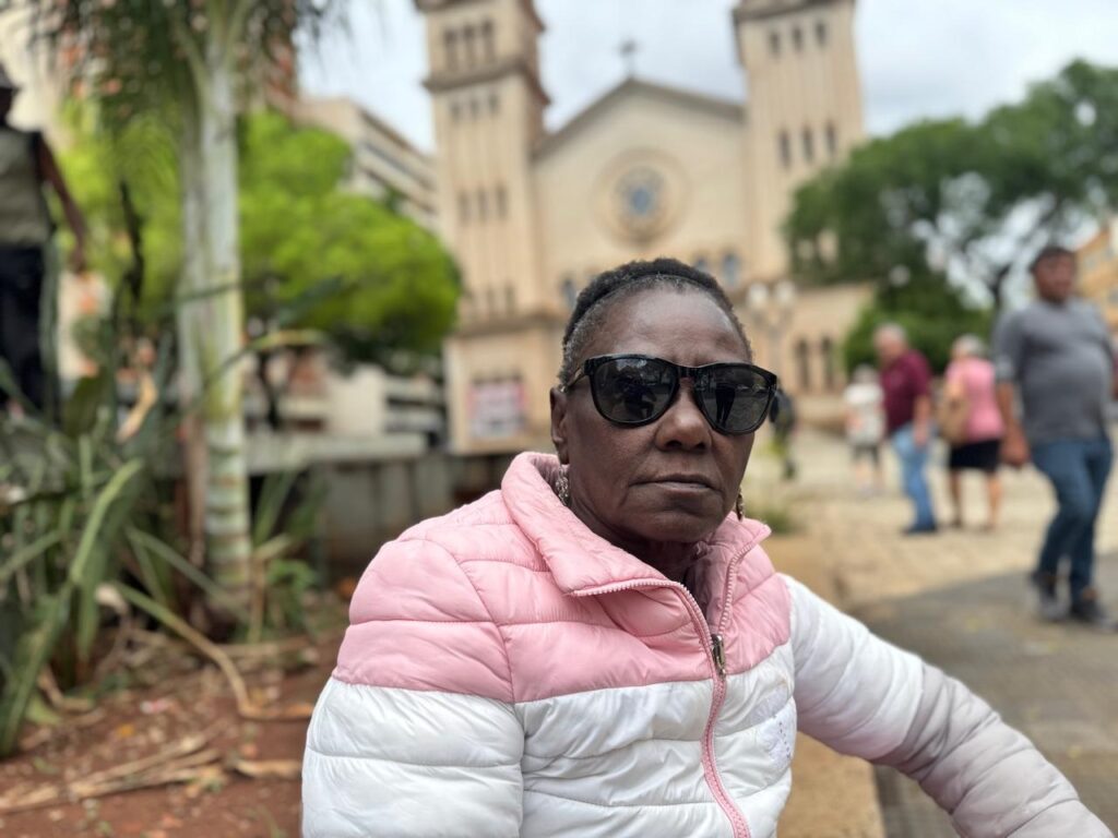 A imagem mostra uma mulher negra de meia-idade, vestindo uma jaqueta acolchoada rosa e branca e óculos escuros. Ela está sentada em um ambiente externo, com uma igreja e outras pessoas desfocadas ao fundo. A iluminação é natural, sugerindo que a foto foi tirada durante o dia. A mulher olha diretamente para a câmera com uma expressão séria. As plantas à esquerda da imagem adicionam um toque de verde ao cenário.