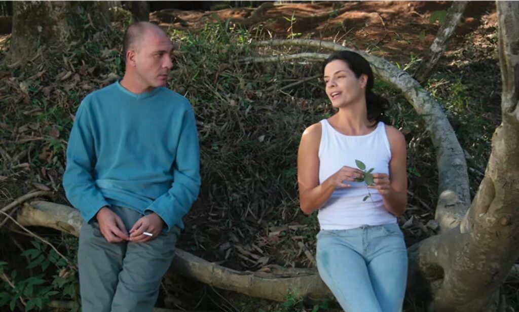 A imagem mostra um homem e uma mulher sentados do lado de fora, em um ambiente natural, possivelmente uma floresta ou parque. O homem, à esquerda, é careca e veste uma blusa azul de manga comprida e calças verdes. Ele está segurando um cigarro entre os dedos. Sua expressão facial sugere que ele está olhando para a mulher. A mulher, à direita, tem cabelos escuros e veste uma blusa branca regata e calças jeans claras. Ela está segurando um pequeno ramo com folhas nas mãos e parece estar falando com o homem, com uma expressão animada. Ambos estão sentados em um tronco caído, e o fundo é composto por vegetação densa, árvores e terra. A luz do sol parece filtrar através das árvores, criando um ambiente natural e relaxante.