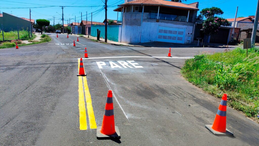 A imagem mostra uma rua em um bairro residencial durante o dia, com céu azul e sol brilhando. No centro da imagem, há a palavra "PARE" pintada em letras grandes e brancas no asfalto, indicando um ponto de parada para veículos. Ao redor da palavra, vários cones de trânsito laranja com faixas refletivas cinzas estão posicionados, possivelmente para sinalizar uma área em obras ou para organizar o fluxo de veículos. As linhas amarelas duplas recém-pintadas delimitam as faixas da via. Casas com telhados de cerâmica e jardins verdes aparecem ao fundo, ao longo da rua. A imagem foi capturada de um ângulo baixo, proporcionando uma visão clara da sinalização no chão e da organização do trânsito naquele local.
