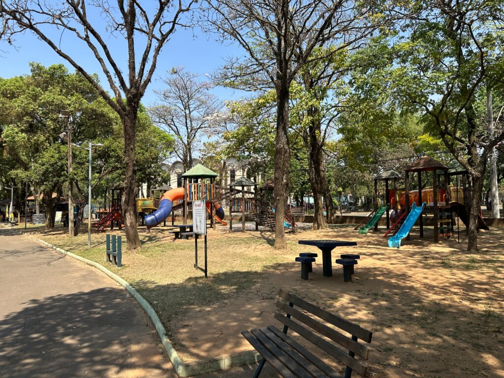A imagem mostra um parque público com uma área de recreação infantil vibrante e colorida, composta por escorregadores, túneis e estruturas para escalada em tons de vermelho, amarelo, azul e verde. Árvores ao redor oferecem sombra e criam um ambiente natural e acolhedor. Há bancos e uma mesa redonda com assentos integrados, ideais para piqueniques ou descanso. O chão é de terra com trechos de grama, e um caminho pavimentado serpenteia pelo espaço.