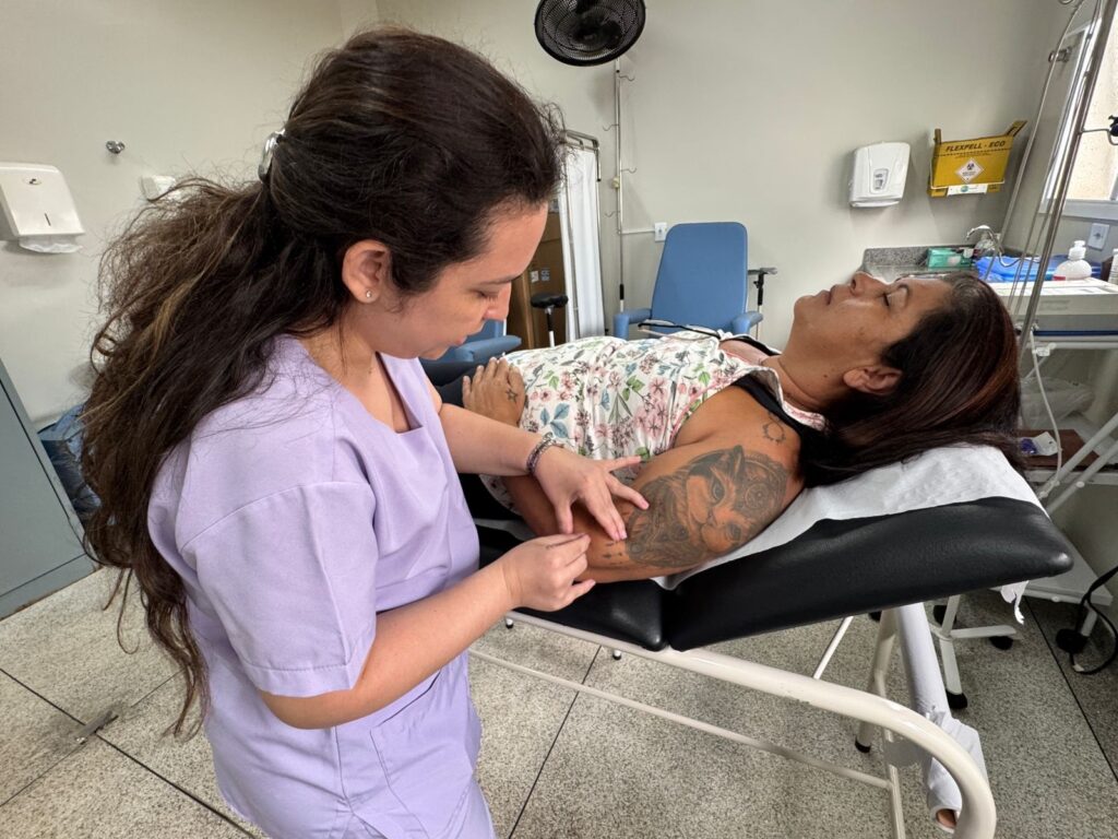 A imagem mostra uma cena médica em que uma profissional de saúde, vestindo um uniforme lilás, está atendendo uma paciente. A profissional tem cabelos longos, escuros e ondulados, presos para trás, e está concentrada no braço da paciente. A paciente está deitada em uma maca, com os olhos fechados e a cabeça inclinada para trás. Ela tem cabelos escuros e usa uma roupa com estampa floral. Seu braço esquerdo, apoiado na maca, apresenta uma tatuagem grande e detalhada de um rosto de felino. As mãos da profissional estão próximas ao braço da paciente, sugerindo que ela pode estar aplicando uma injeção ou verificando uma veia. O ambiente parece ser uma clínica ou hospital, com equipamentos médicos ao fundo, incluindo uma cadeira azul, um carrinho com materiais e um dispensador na parede. A iluminação é clara e típica de ambientes médicos. A imagem destaca o cuidado e a atenção na interação entre as duas pessoas.