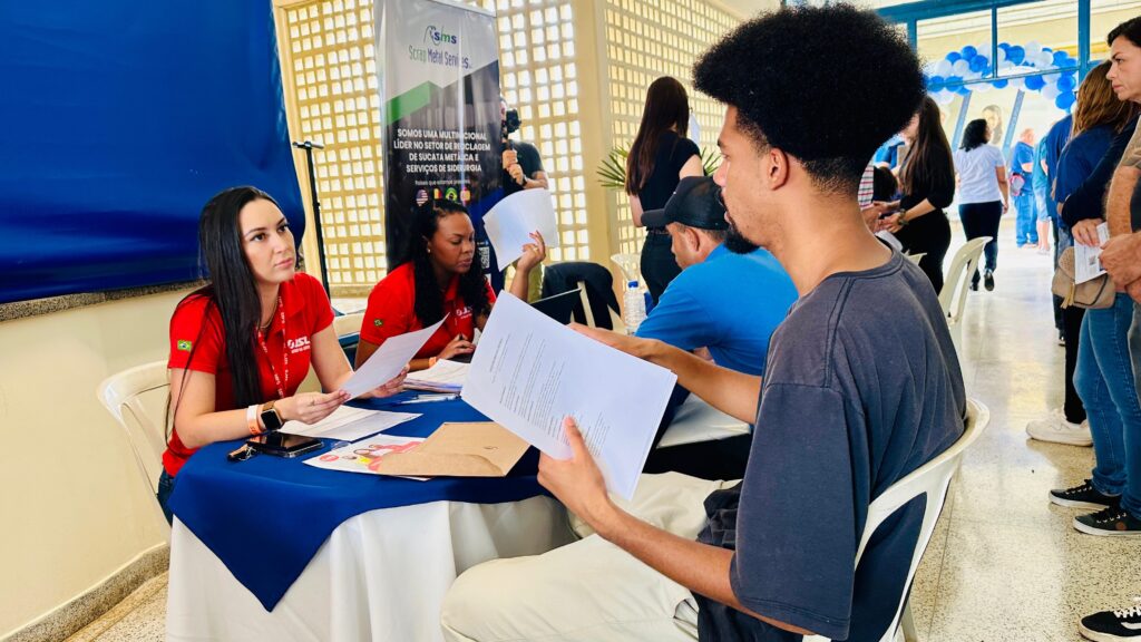 A imagem mostra um evento ao ar livre, possivelmente uma feira de empregos ou um evento de recrutamento. Há várias pessoas em pé e sentadas, interagindo em torno de mesas. No primeiro plano, à esquerda, uma mulher com uma camisa vermelha da "GISL" (com uma bandeira do Brasil na manga) está sentada a uma mesa coberta com uma toalha azul. Ela segura um papel e parece estar conversando com alguém fora do quadro. Ao lado dela, outra mulher, também com uma camisa vermelha semelhante, está sentada e olhando para um papel. À direita, um homem com cabelo afro e uma camiseta cinza está sentado, segurando um papel e olhando para ele, possivelmente em discussão com outra pessoa. Ao fundo, há um banner com o logotipo "sms Scrap Metal Services" e texto em português que parece indicar que a empresa é uma multinacional líder em reciclagem de sucata metálica e serviços de siderurgia. Há também balões azuis e brancos decorando a área, sugerindo um ambiente festivo ou de evento. Outras pessoas estão em fila ou conversando ao fundo. A iluminação sugere que o evento está ocorrendo durante o dia.