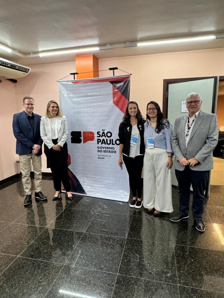Um grupo de cinco pessoas está posicionado em frente a um banner com o logo do Governo do Estado de São Paulo e a inscrição "SÃO PAULO GOVERNO DO ESTADO Secretaria da Saúde". Da esquerda para a direita, vemos um homem de terno azul escuro, uma mulher com um blazer branco e calça preta, outra mulher com um blazer preto e calça preta, uma terceira mulher com uma blusa listrada e calça branca, e um homem de terno cinza claro. Todos parecem estar em um evento ou reunião, e as três mulheres usam crachás. O piso é escuro e brilhante, e há iluminação no teto.