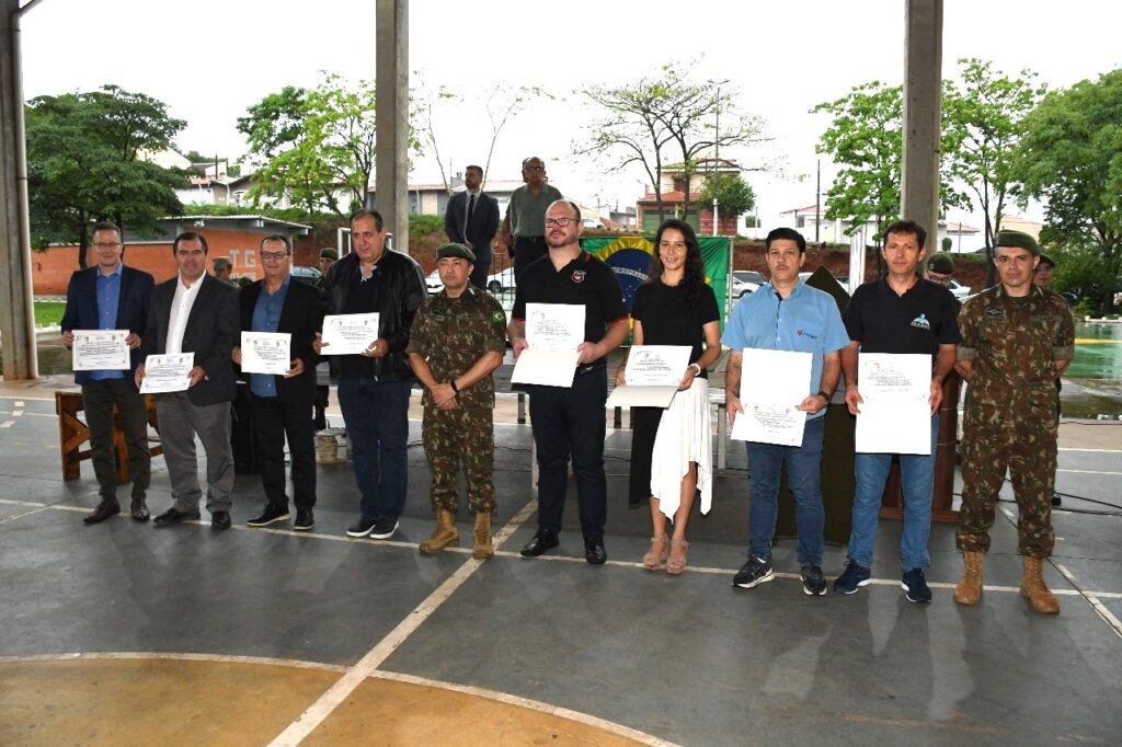 A imagem mostra um grupo de pessoas em pé, alinhadas, recebendo certificados. Há uma bandeira do Brasil ao fundo, e algumas árvores e prédios. As pessoas estão usando roupas variadas, algumas em trajes civis e outras em uniformes militares. A maioria das pessoas está segurando um certificado em mãos. O evento parece ser uma cerimônia de entrega de diplomas ou reconhecimentos.