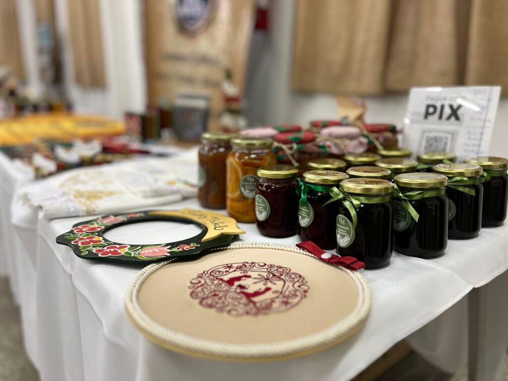 A imagem exibe uma mesa coberta com uma toalha branca, onde estão dispostos diversos produtos artesanais. Em destaque, há uma fileira de potes de vidro com tampas douradas, contendo geleias de diferentes cores e sabores, decorados com fitas coloridas (vermelha, verde e rosa) e rótulos circulares com um monograma "M" e o nome "Ana Moreira". Atrás dos potes, mais geleias estão empilhadas. À frente dos potes, repousam dois itens decorativos. Um deles é um porta-retrato em formato irregular, pintado de preto e decorado com flores vermelhas e rosas, com detalhes em verde e amarelo. Ao lado e ligeiramente abaixo, encontra-se um bastidor de bordado com um tecido bege e um bordado em tom de vinho, representando uma figura feminina estilizada dentro de um desenho intrincado. Ao fundo, desfocados, visualizam-se mais produtos, como tecidos estampados e sacolas de papel kraft com logotipos, sugerindo um ambiente de feira de artesanato ou venda de produtos locais. Há também um cartaz com a palavra "PIX" e um QR code, indicando que pagamentos podem ser feitos por esse método. A iluminação suave contribui para uma atmosfera acolhedora e artesanal.