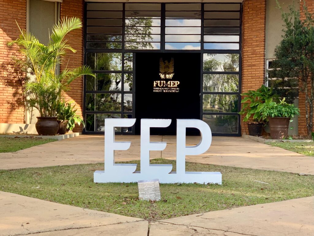 A imagem mostra a entrada de um prédio com uma fachada de tijolos e uma grande porta de vidro. Em primeiro plano, há um letreiro branco com as letras "EEP" em relevo, posicionado sobre um gramado. Ao fundo, atrás do letreiro, está a porta de vidro, que reflete o céu e a vegetação ao redor. Acima da porta, há um painel preto com o logotipo dourado da "FUMEP - FUNDAÇÃO MUNICIPAL DE ENSINO DE PIRACICABA". À esquerda da porta, há um vaso grande com uma planta alta e frondosa. À direita, há vasos menores com plantas verdes. O caminho em frente ao prédio é pavimentado com pedras claras.