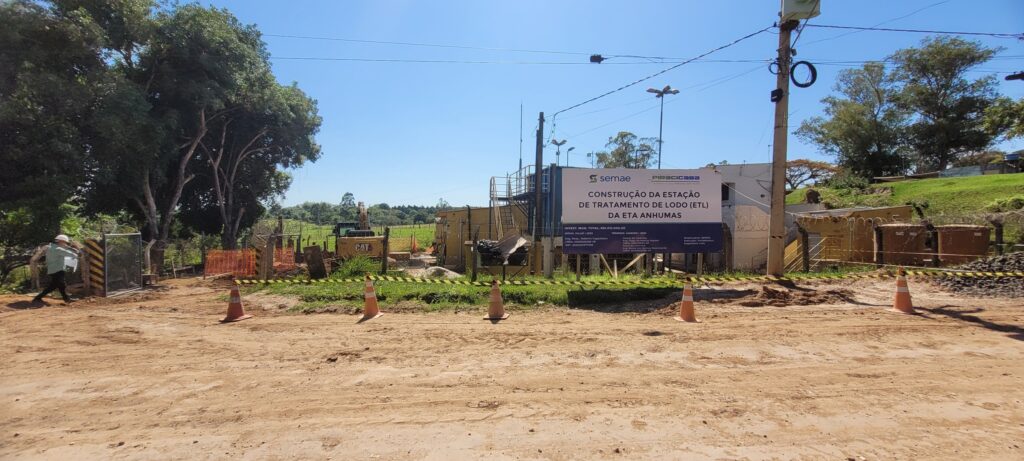 A imagem mostra o canteiro de obras para a construção de uma Estação de Tratamento de Lodo (ETL) da ETA Anhumas. Um grande banner informa sobre o projeto, com os logotipos da Semae e Piracicaba. Ao fundo, há equipamentos de construção, como uma escavadeira, e estruturas temporárias. O local está cercado por grades e fitas de isolamento, com cones de trânsito laranja e preto sinalizando o acesso. Um homem vestindo capacete e colete de segurança caminha na entrada do canteiro. O céu está claro e azul, indicando um dia ensolarado. Há árvores frondosas e vegetação ao redor, com uma área de gramado em elevação ao fundo.