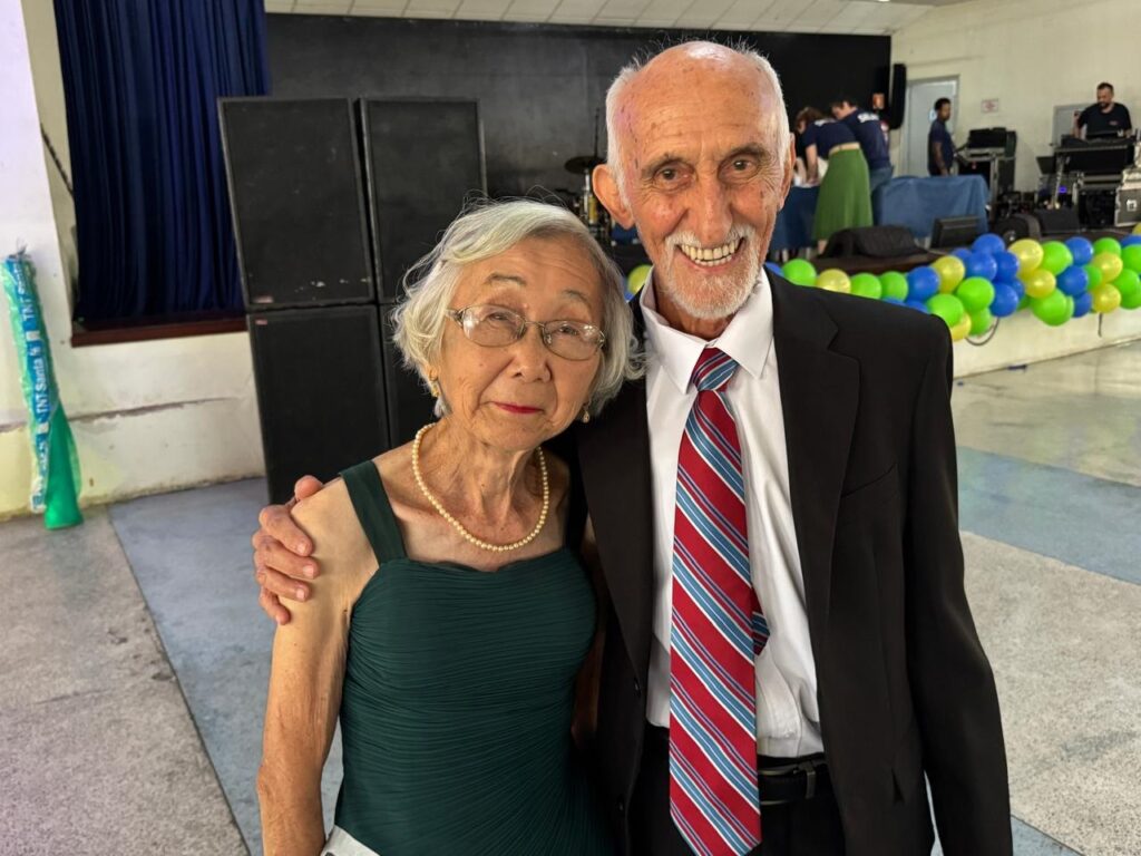 A imagem mostra um casal de idosos sorrindo para a câmera. O homem, à direita, usa um terno preto, camisa branca e gravata listrada em vermelho, branco e azul. A mulher, à esquerda, veste um vestido verde escuro e um colar de pérolas. Ambos parecem estar em um evento, com balões azuis e verdes e uma banda ao fundo.