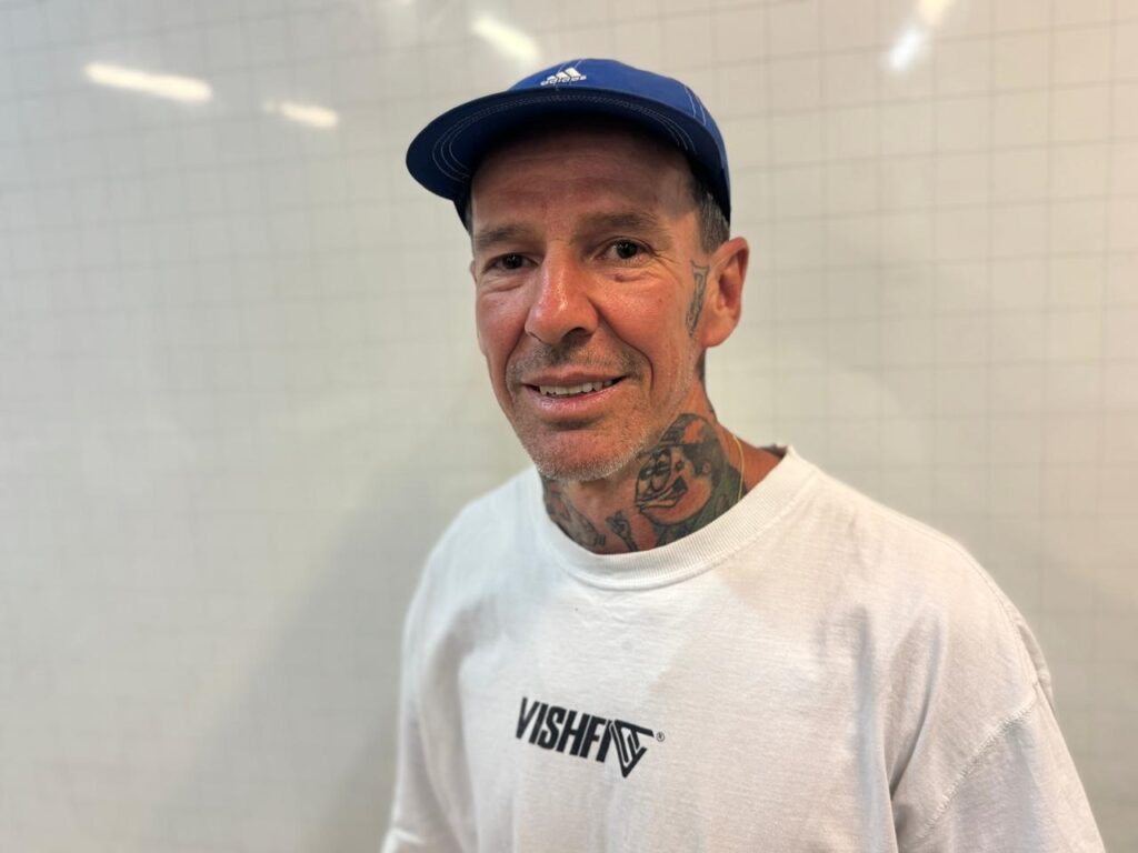 A imagem mostra um homem em um retrato médio, com tatuagens visíveis no pescoço e no peito. Ele usa um boné azul e uma camiseta branca com um logotipo preto que diz "VISH". O homem está olhando para a câmera com um leve sorriso. O fundo é uma parede branca lisa com um padrão sutil, sugerindo um ambiente interno, como um escritório ou estúdio. A iluminação é clara e uniforme, destacando o rosto e a parte superior do corpo do homem. A composição é simples, focando no sujeito centralizado na imagem.