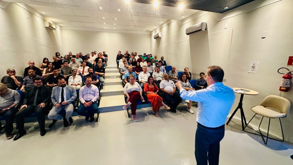 A imagem mostra uma sala de conferências com uma audiência sentada em degraus, voltada para um palestrante em pé. O palestrante, vestindo uma camisa azul clara e calças escuras, está de costas para a câmera, gesticulando com as mãos enquanto fala. A audiência é composta por homens e mulheres de diversas idades, a maioria vestida de forma casual ou profissional. As paredes da sala são claras, com iluminação embutida no teto. Há dois aparelhos de ar condicionado visíveis nas paredes. No lado direito da imagem, há um extintor de incêndio vermelho pendurado na parede e uma cadeira bege com encosto e assento estofados ao lado de uma pequena mesa redonda com um tripé. Uma folha de papel com um código QR está afixada na parede atrás da mesa. O piso da sala é de cor clara e lisa.