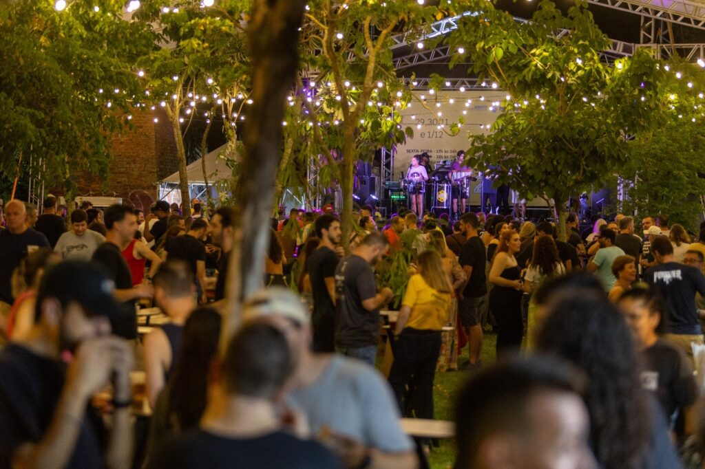 A imagem retrata um evento ao ar livre, possivelmente um festival ou show, à noite. A cena é iluminada por uma série de luzes penduradas em árvores, criando uma atmosfera festiva e aconchegante. Ao fundo, um palco com músicos tocando e uma banda se apresenta, com luzes coloridas iluminando o espaço. Uma multidão de pessoas está reunida, algumas em pé e outras sentadas, interagindo e aproveitando o momento. A vegetação, com árvores frondosas e iluminadas, adiciona um toque natural ao ambiente. A sensação geral é de alegria e celebração.
