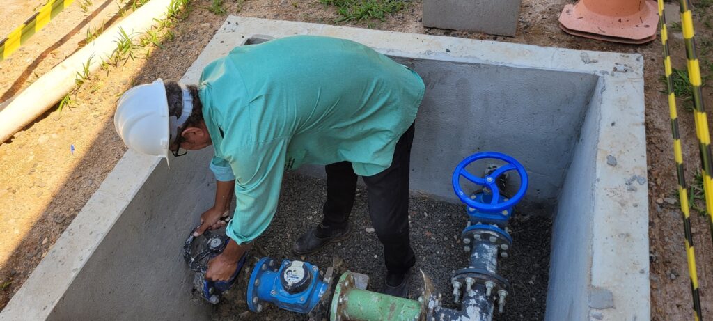 A imagem mostra um trabalhador usando um capacete branco e uma camisa verde, agachado dentro de uma estrutura de concreto. Ele está mexendo em equipamentos de encanamento, incluindo um medidor de água azul e uma válvula azul. Ao lado, há uma tubulação verde e uma seção de tubulação preta com uma válvula azul com uma roda de manobra. O ambiente é externo, com terra, cascalho e grama visíveis. Há também uma fita de segurança amarela e preta e um cone laranja em segundo plano.