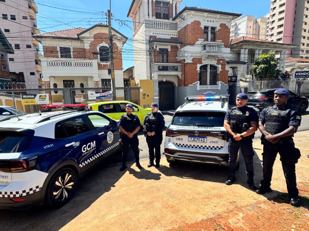 A imagem mostra quatro agentes da Guarda Civil Municipal (GCM) em frente a duas viaturas, estacionadas em uma rua com edifícios residenciais ao fundo. As viaturas são uma azul e branca com a inscrição "GCM PIRACICABA" e uma branca com a inscrição "PATRULHA MARIA DA PENHA". Os agentes estão uniformizados, usando coletes táticos e boinas. Alguns edifícios ao fundo possuem placas de "ALUGA" e "VENDO". O dia está ensolarado, com céu azul e poucas nuvens.