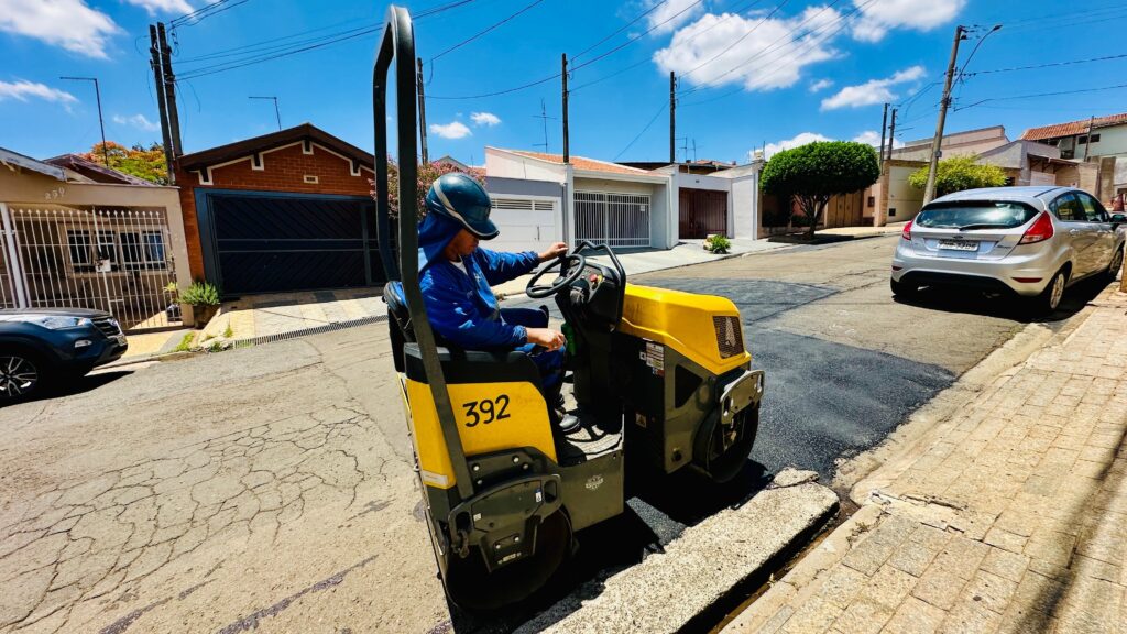 O homem está operando um rolo compactador amarelo na rua. Ele está usando um macacão azul e um capacete azul. O rolo compactador está esmagando o asfalto fresco na rua. Há casas e carros ao fundo. O dia está ensolarado com algumas nuvens no céu.