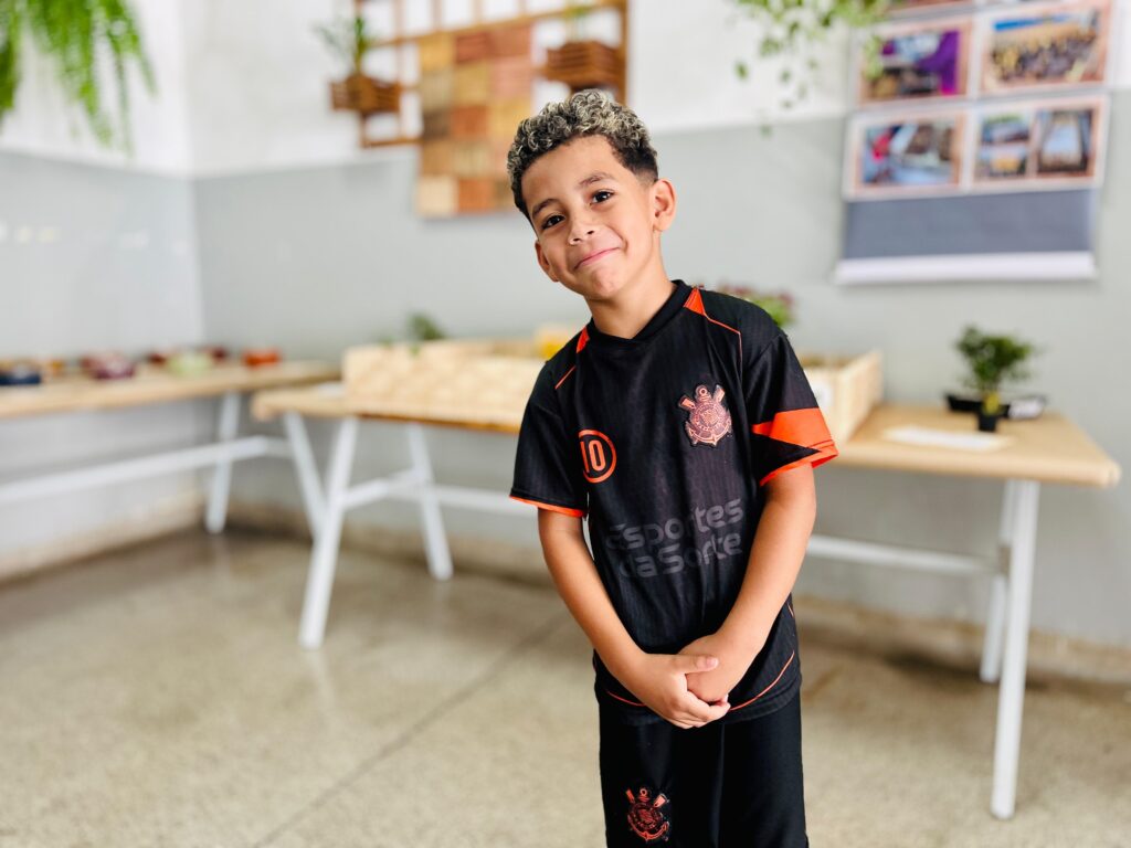 A imagem mostra um menino sorridente em pé em uma sala, possivelmente uma sala de aula ou um espaço de atividades. Ele está vestindo um uniforme de futebol preto com detalhes em laranja, que inclui uma camisa com o número 10 e um brasão, e shorts com o mesmo brasão. O cabelo do menino é encaracolado e descolorido nas pontas. Ao fundo, há mesas com objetos dispostos sobre elas, como bandejas com plantas ou materiais, e prateleiras com decorações e fotos. A iluminação é clara, sugerindo um ambiente interno bem iluminado. O piso é de concreto polido. O foco principal da imagem é o menino, que olha diretamente para a câmera com uma expressão amigável.