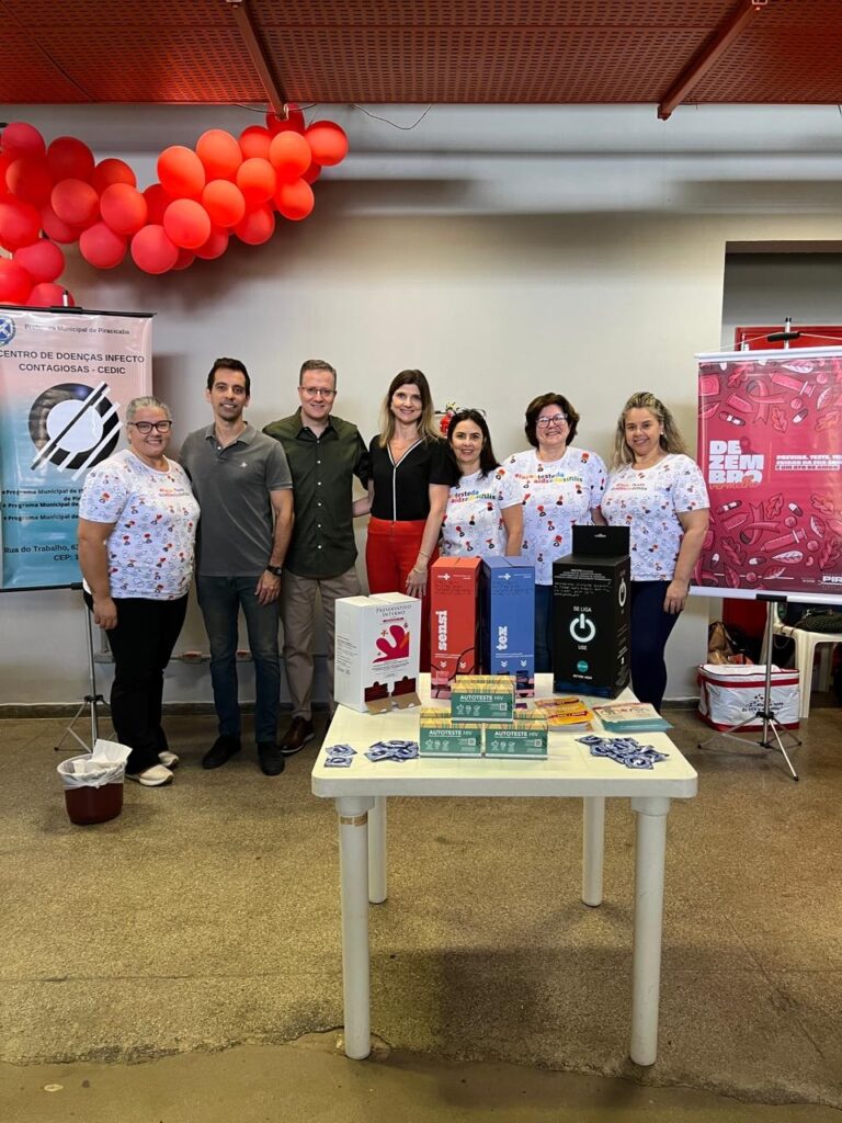 A imagem mostra um grupo de sete pessoas em pé atrás de uma mesa com produtos. O grupo é composto por cinco mulheres e dois homens, todos vestindo roupas casuais. As mulheres usam camisetas brancas com estampas coloridas, e algumas delas têm o texto "tested aids/syphilis" em suas camisetas. Os homens vestem camisas de cores diferentes. Na mesa, há vários produtos, incluindo caixas de testes de HIV (AUTOTESTE HIV), preservativos de diferentes marcas (Sensi, Tez), e uma caixa branca com a inscrição "PRESERVATIVO INTERNO". Há também um cartaz ao fundo com a frase "DEZEMBRO VERMELHO" e informações sobre prevenção de HIV/AIDS. O ambiente parece ser um evento de conscientização sobre saúde, possivelmente relacionado à AIDS, com balões vermelhos decorando o teto.