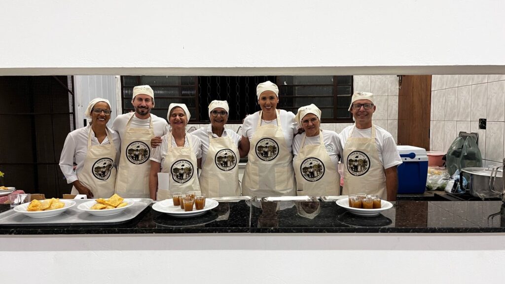 Sete pessoas, cinco mulheres e dois homem, estão em pé atrás de um balcão, todos vestindo aventais brancos com um logotipo circular e chapéus de chef. O homem, no centro, está com um sorriso largo e usa óculos. As mulheres ao seu redor também sorriem, algumas com óculos. Na frente deles, sobre o balcão, há pratos com comida. Em duas bandejas retangulares, há vários pastéis dourados fritos. Em um prato redondo à esquerda, há mais pastéis. Em um prato redondo à direita, há vários copinhos de shot com um doce líquido âmbar, possivelmente caramelo ou doce de leite. O fundo parece ser uma cozinha ou área de preparação de alimentos, com azulejos na parede e alguns equipamentos visíveis, como uma caixa térmica azul e panelas. A iluminação é um pouco escura, dando um tom acolhedor à cena.