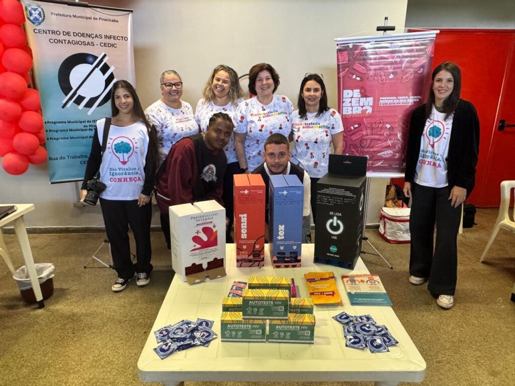 A imagem mostra um grupo de pessoas em pé atrás de uma mesa cheia de materiais de saúde sexual, incluindo preservativos, testes rápidos de HIV e folhetos informativos. Há também caixas com a marca "Sensi" e "Tex", e uma caixa maior rotulada como "Preservativo Interno". Ao fundo, um banner com a inscrição "Dezembro Vermelho" sugere que o evento está relacionado à conscientização sobre HIV/AIDS. Várias pessoas no grupo usam camisetas com mensagens relacionadas à saúde e prevenção. O ambiente parece ser um evento de saúde pública, possivelmente em uma prefeitura ou centro de saúde local, dada a presença de um banner com o brasão da Prefeitura Municipal de Piracicaba.