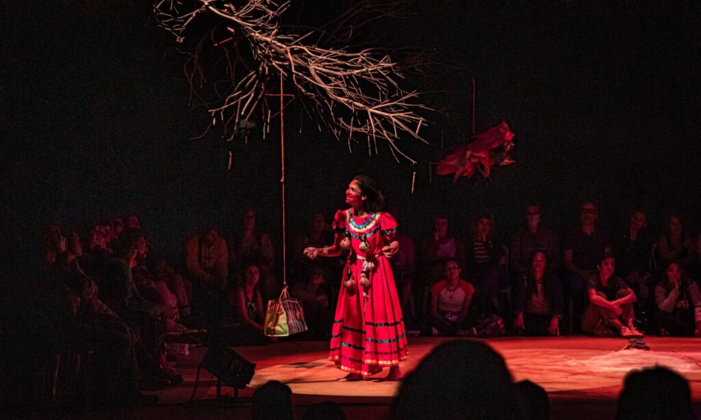 A imagem retrata uma cena teatral com iluminação dramática, focada em uma atriz principal. Ela está no centro do palco, vestindo um vibrante vestido vermelho com detalhes em azul e dourado, e seu rosto está pintado com maquiagem vermelha. A atriz está em uma pose expressiva, com os braços ligeiramente abertos, como se estivesse no meio de uma performance. Acima dela, um galho seco e retorcido se projeta do teto, com alguns fios finos pendurados. À esquerda da atriz, uma bolsa com listras coloridas está suspensa por um fio. À direita, em um plano mais distante, um objeto vermelho e volumoso, possivelmente uma flor estilizada ou um elemento de figurino, também está pendurado. O palco é iluminado por uma luz vermelha intensa, que cria uma atmosfera sombria e misteriosa. Ao fundo, sentada em semicírculo, está a plateia, cujos rostos e figuras são parcialmente visíveis, mas desfocados pela escuridão e pela distância. A plateia parece atenta à performance, com alguns espectadores visíveis em primeiro plano, também em silhueta. A imagem captura um momento de intensa atuação e a atmosfera envolvente de uma apresentação teatral.