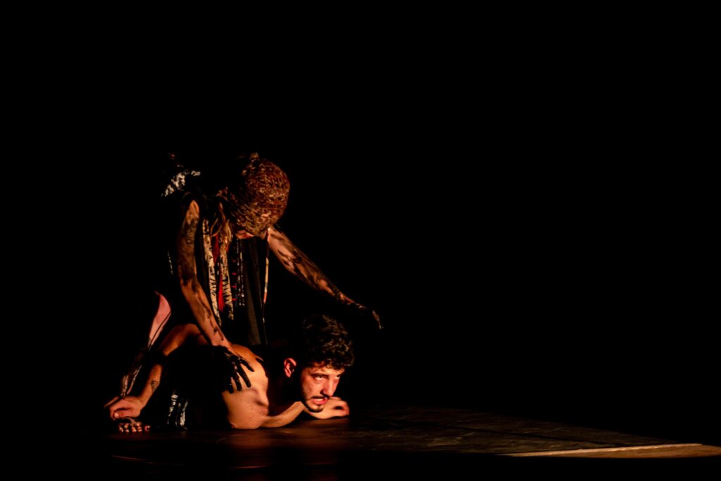 Na imagem, dois indivíduos estão em cena sobre um palco escuro, iluminados por uma luz dramática que destaca a intensidade do momento. Um deles está deitado de bruços no chão, com o corpo tenso, transmitindo sofrimento ou vulnerabilidade. Atrás dele, uma figura imponente, com pintura corporal e traje elaborado, posiciona as mãos sobre suas costas, sugerindo um gesto ritualístico, de domínio ou transformação. O contraste entre a luz focada e o fundo sombrio acentua o clima teatral e emocional.