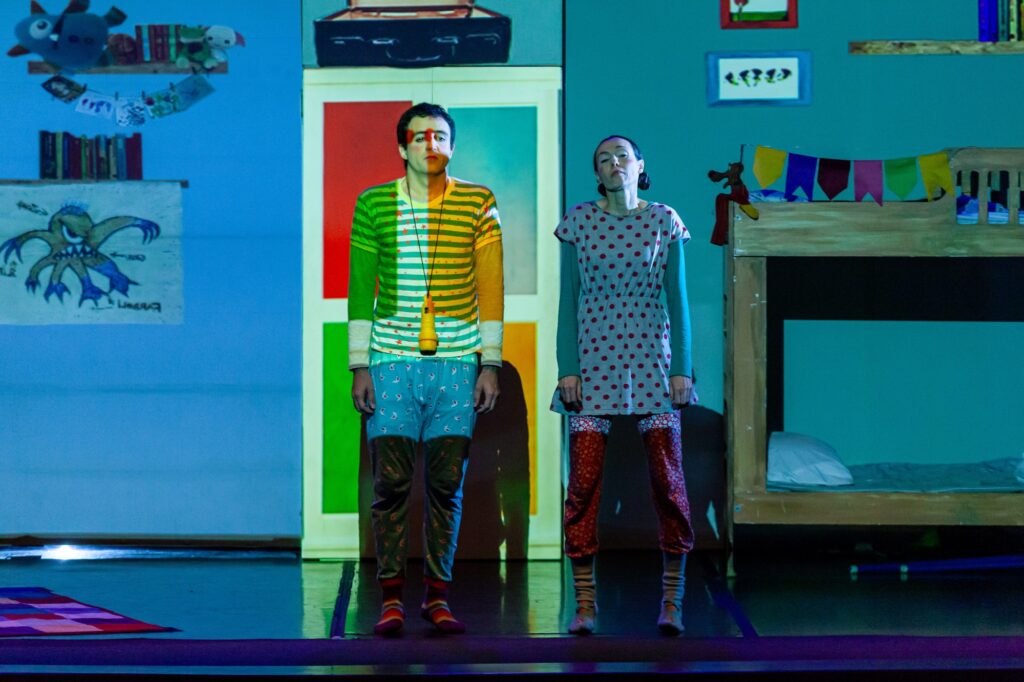 Na imagem, duas pessoas estão em pé diante de uma porta intensamente iluminada, em um ambiente que lembra o quarto de uma criança ou um cenário teatral inspirado nesse universo. A luz projeta faixas coloridas sobre seus rostos e roupas, criando um efeito surreal e mágico. À esquerda, alguém veste camiseta listrada e shorts com estrelas; à direita, outra figura usa vestido de bolinhas e meias estampadas. Ambos têm rostos pintados, reforçando o clima de fantasia e performance. Ao fundo, há um beliche decorado com bandeirolas, desenhos infantis nas paredes — como um polvo — e um tapete colorido no chão, compondo um espaço lúdico e imaginativo.