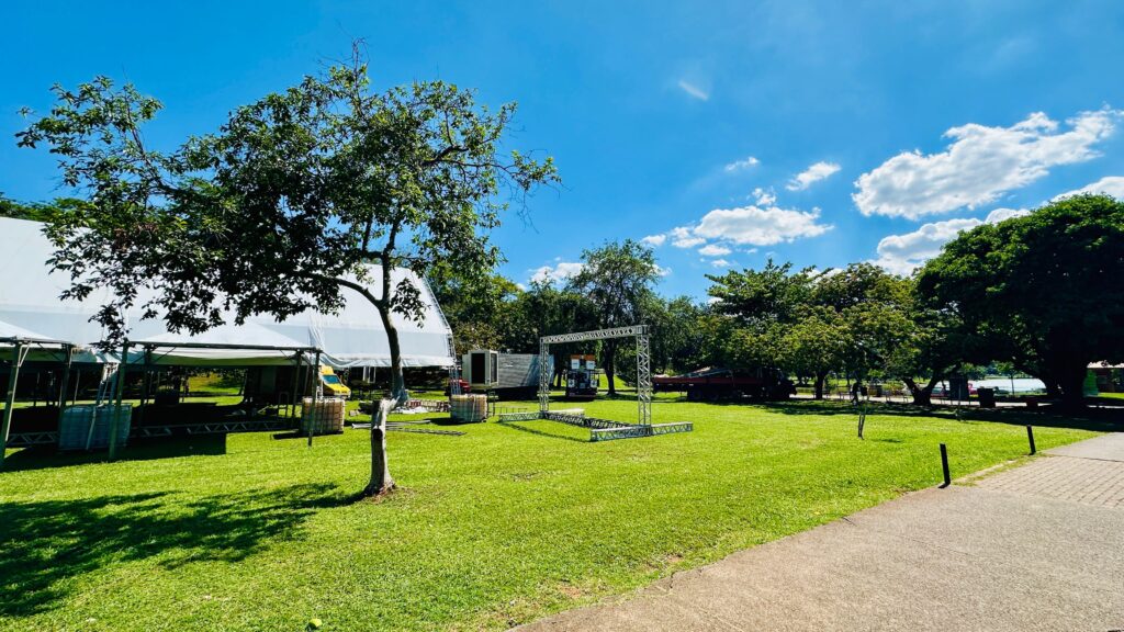A imagem mostra um gramado verde sob um céu azul claro com algumas nuvens brancas. Há uma grande tenda branca montada à esquerda, com uma estrutura de metal visível por baixo. Ao lado da tenda, há um pequeno caminhão amarelo. No centro da imagem, uma estrutura de metal em forma de arco está montada no gramado, com equipamentos de som e iluminação visíveis. Há também um reboque vermelho e outras estruturas de metal espalhadas pela área. Árvores frondosas com folhas verdes cobrem o fundo da imagem, com uma grande árvore em primeiro plano à esquerda. Ao fundo, pode-se ver um lago ou corpo de água. O gramado está bem cuidado e a cena parece ser de um evento ao ar livre que está sendo preparado.