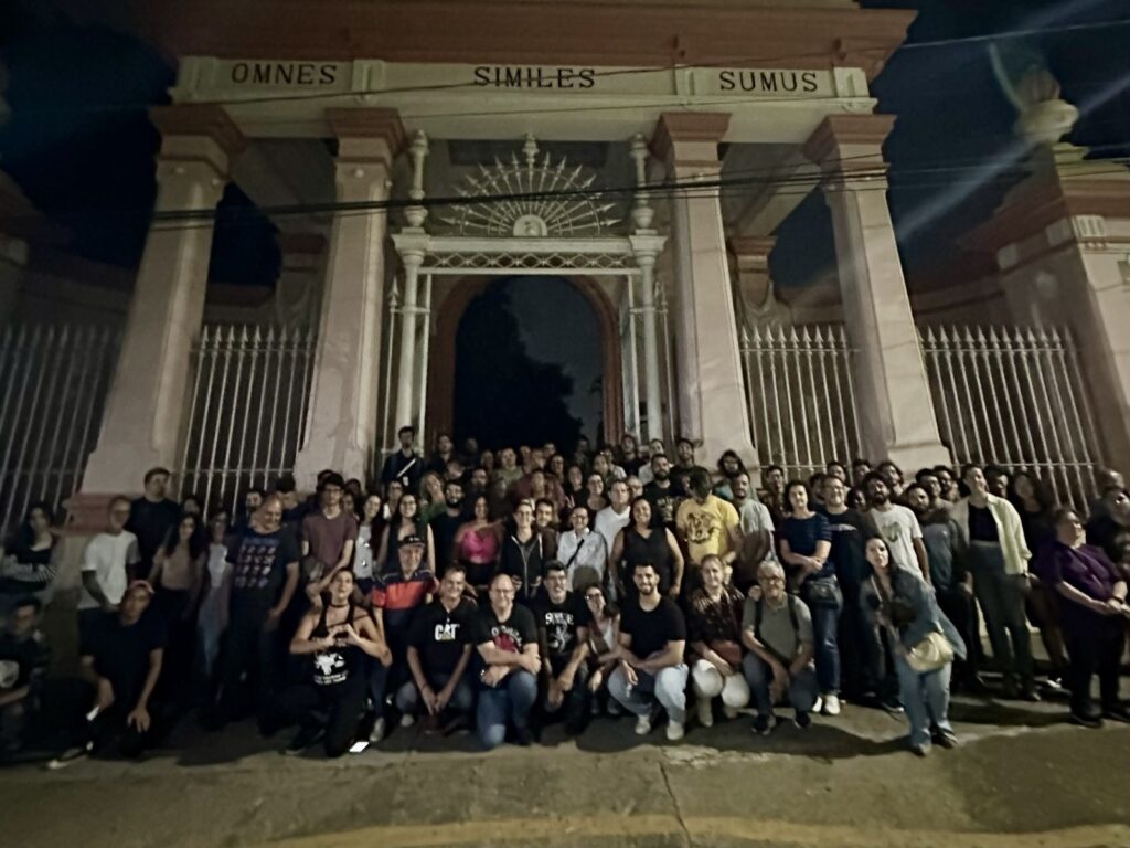 A imagem mostra um grande grupo de pessoas reunidas em frente a uma estrutura arquitetônica imponente, que parece ser a entrada de um edifício histórico ou monumento. A iluminação é noturna, com luzes artificiais que iluminam a fachada e as pessoas. A estrutura principal é composta por um arco central com grades ornamentadas, ladeado por colunas robustas e arcos menores. Acima do arco principal, há uma inscrição em letras maiúsculas: "OMNES SIMILES SUMUS". As cores predominantes na arquitetura são o rosa claro e o branco, com detalhes em tons mais escuros. O grupo de pessoas, composto por dezenas de indivíduos de diversas idades e gêneros, está posicionado em frente à entrada, alguns em pé e outros agachados ou sentados no chão. Eles parecem estar posando para uma fotografia, com muitos olhando para a câmera. As roupas variam, mas muitos usam roupas casuais.