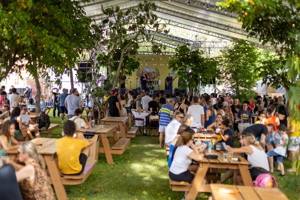 A imagem retrata um festival ao ar livre, possivelmente um show ou evento musical, sob uma tenda com estrutura metálica e cobertura transparente. Há um palco montado ao fundo, com uma banda se apresentando. A área frontal e intermediária está repleta de pessoas sentadas em mesas de piquenique de madeira, interagindo e desfrutando do evento. O gramado verde serve de chão, e árvores frondosas com iluminação decorativa (pisca-piscas) criam um ambiente agradável e festivo. A multidão é diversificada em termos de idade e vestimenta, indicando um evento popular e familiar. A luz natural, combinada com a iluminação artificial, sugere que o evento ocorre durante o dia ou no início da noite.