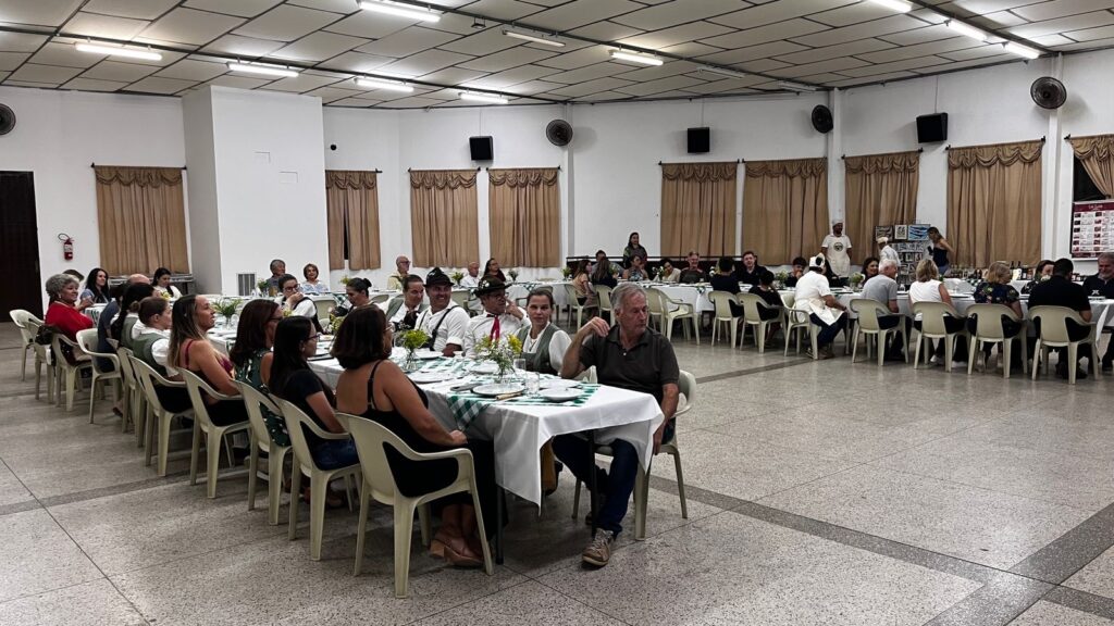 A imagem mostra um salão de festas com várias mesas postas para um jantar ou evento. Há muitos convidados sentados às mesas, conversando e interagindo. Algumas pessoas usam trajes típicos, sugerindo uma celebração cultural. As mesas estão cobertas com toalhas brancas e xadrez, com pratos, copos e arranjos florais. Ao fundo, observa-se uma área onde pessoas parecem estar servindo ou organizando o evento. As paredes são claras, com cortinas marrons e iluminação embutida no teto. O ambiente parece ser informal, mas organizado para um encontro social.
