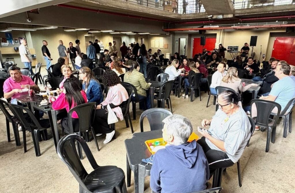 A imagem retrata um ambiente interno com várias pessoas sentadas em mesas e cadeiras. Ao fundo, há uma estrutura de metal no teto e paredes com algumas quadros e cartazes. Há também uma banda tocando ao fundo, com instrumentos como violão e teclado. As pessoas parecem estar em um momento de confraternização ou evento, com algumas interagindo e outras focadas em seus celulares. A iluminação é artificial, vinda de luminárias no teto.