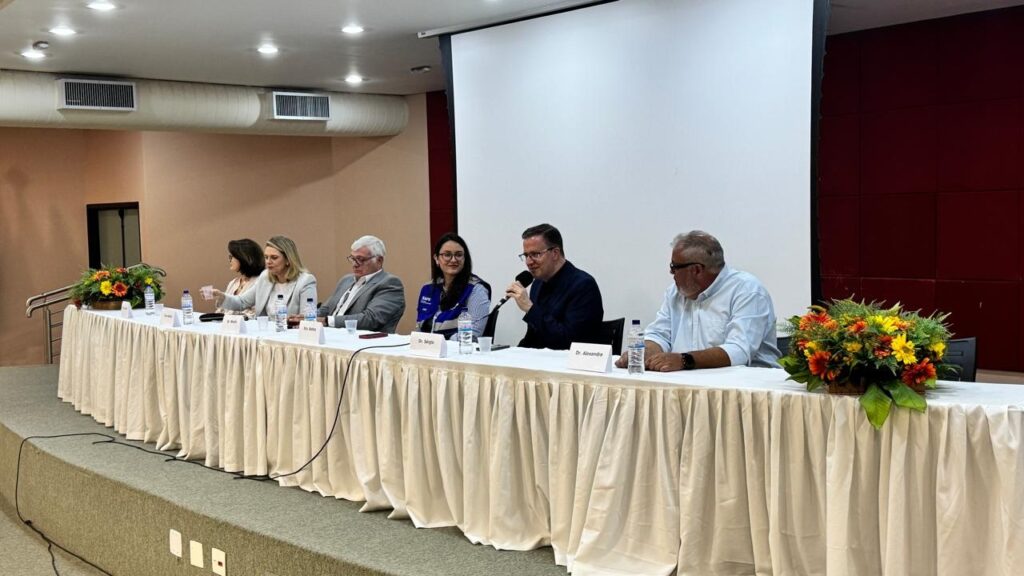 A imagem mostra um painel de discussão com seis pessoas sentadas a uma longa mesa coberta por uma toalha branca com drapeado. Todos estão voltados para a frente, com exceção de um homem no extremo direito que está virado para a esquerda. À esquerda, há um arranjo de flores em tons de laranja, amarelo e vermelho. As pessoas sentadas na mesa parecem estar em um evento formal, pois estão vestidas de forma profissional. Há garrafas de água e copos sobre a mesa, e um microfone está posicionado em frente a um dos palestrantes. Uma tela branca está visível ao fundo, sugerindo que o evento pode incluir uma apresentação. A iluminação é suave, criando uma atmosfera profissional e acolhedora.