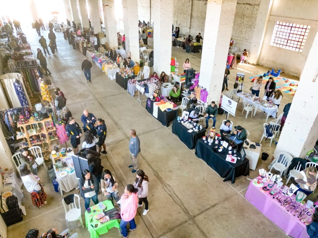 A imagem mostra um mercado interno movimentado, com muitas pessoas circulando entre as barracas de venda. O espaço parece ser um galpão industrial antigo, com colunas de tijolos aparentes e um teto alto com estrutura metálica. As barracas oferecem uma variedade de produtos, incluindo alimentos, artesanato e roupas. Em primeiro plano, vemos um casal mais velho passeando e, à sua esquerda, uma barraca com diversos doces e bolos em exposição. Mais adiante, outras barracas exibem pães, doces e outros produtos alimentícios, com vendedores atendendo aos clientes. Pessoas de diversas idades e estilos circulam pelo local, algumas observando os produtos, outras conversando e comprando. Ao fundo, a perspectiva se estende por um longo corredor, onde mais barracas e pessoas compõem a cena. A iluminação é uma mistura de luz natural que entra pelas janelas altas e luzes artificiais do teto. O ambiente geral é de um evento popular e animado, possivelmente uma feira de artesanato ou de produtos locais.