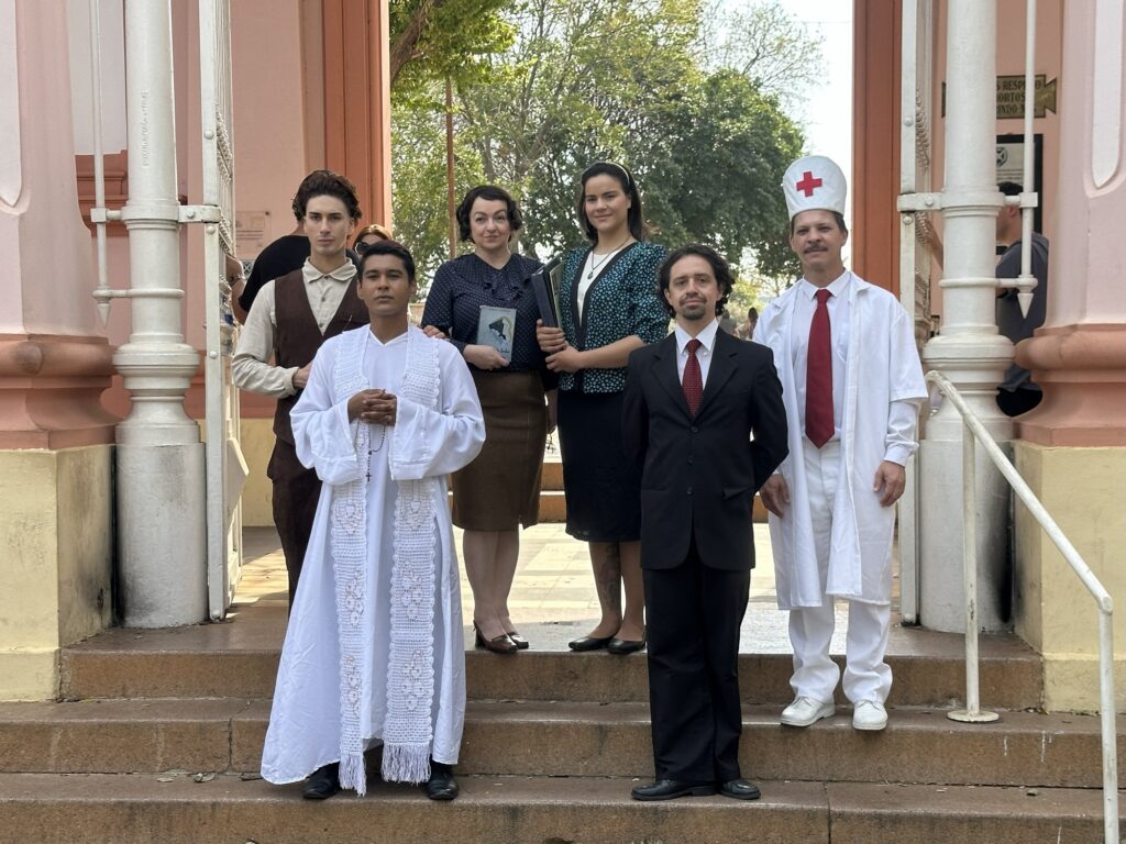 Na imagem, um grupo de seis pessoas está posicionado em degraus de uma edificação com colunas e detalhes arquitetônicos em tons pastéis. Do lado esquerdo, um jovem veste uma camisa bege clara e um colete marrom, enquanto ao seu lado, um homem em pé ostenta um hábito religioso branco com detalhes em renda e um rosário, com as mãos em prece. Em seguida, uma mulher com cabelo escuro preso, veste uma blusa estampada e uma saia marrom, segurando um livro. Ao seu lado, outra mulher, com cabelo mais claro, usa uma blusa com estampa de bolinhas e um blazer escuro, também segurando um livro. No centro, um homem veste um terno preto com gravata vermelha. Finalizando o grupo, à direita, um homem com bigode usa um jaleco branco, uma gravata vermelha e um chapéu de enfermeiro com uma cruz vermelha, sugerindo ser um profissional da saúde. O fundo mostra árvores e a fachada de uma construção.