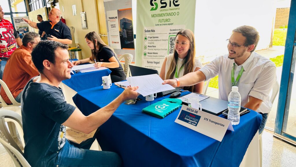 A imagem retrata um evento de recrutamento, possivelmente uma feira de empregos, onde pessoas interagem em mesas com toalhas azuis. Em primeiro plano, um homem de camiseta escura e jeans entrega papéis a uma mulher sentada atrás de uma mesa, que usa óculos e uma blusa branca com um crachá verde. Ao lado dela, um homem de camisa branca e óculos sorri, também com um crachá verde. A mesa possui um laptop, uma garrafa de água e um folder verde com um símbolo de reciclagem. Ao fundo, vemos outras mesas com pessoas conversando e um banner da empresa "STE Logística e Transportes", que promove a ideia de "Movimentando o Futuro" e convida para se juntar à equipe. O banner também detalha informações sobre a empresa, como o que fazem, seu foco, valores e que há vagas disponíveis, incentivando a diversidade e inclusão. Outro banner menor, com o título "TRABALHE CONOSCO", também está visível. Há também outras pessoas em segundo plano, indicando um ambiente de evento movimentado.