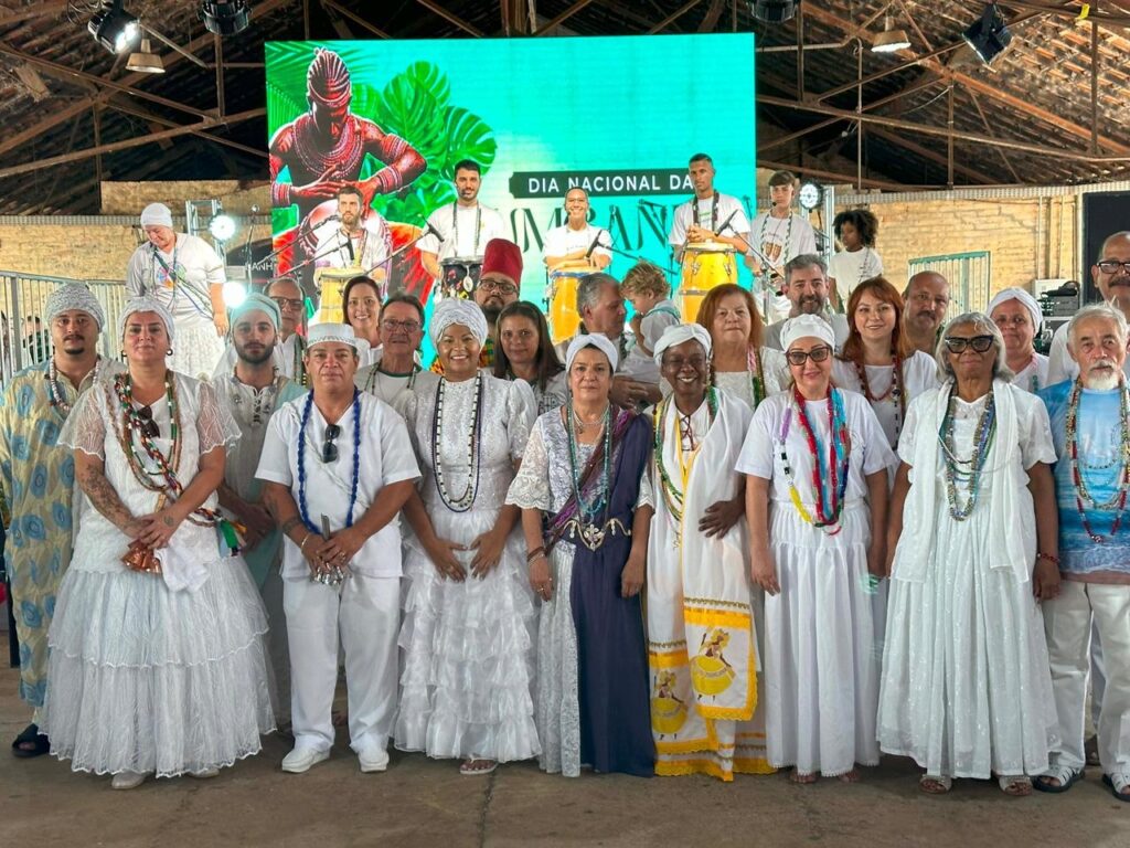 A imagem mostra um grupo de pessoas vestidas com trajes brancos e coloridos, adornadas com colares e fitas. Elas estão em pé e posam para a foto em um ambiente que parece ser um palco ou um evento cultural, com um grande telão ao fundo exibindo uma imagem estilizada de um guerreiro e plantas tropicais, além de um texto que parece ser "DIA NACIONAL DA" UMBANDA" A maioria das pessoas usa turbantes ou lenços na cabeça, e os trajes brancos variam em estilo, alguns com babados e rendas, enquanto outros são mais simples. Os colares e fitas adicionam um toque de cor vibrante aos trajes, com tons de azul, verde, amarelo, vermelho e roxo. A disposição do grupo sugere uma celebração ou cerimônia, com pessoas de diferentes idades e gêneros presentes.