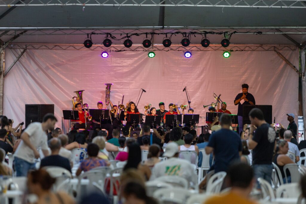 A imagem mostra uma banda de metais se apresentando em um palco externo, com uma cortina branca ao fundo e luzes coloridas acima. Os músicos estão sentados em cadeiras, tocando seus instrumentos, que incluem trombones, tubas e trompetes. Há um cantor no centro do palco, segurando um microfone. Na frente do palco, uma plateia está sentada em cadeiras brancas, assistindo à apresentação. Algumas pessoas na plateia estão tirando fotos com seus celulares.