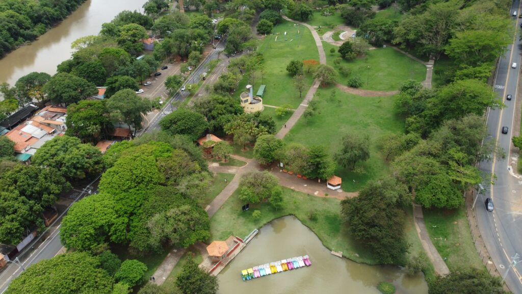 A imagem aérea revela um parque vibrante, repleto de verde exuberante e um rio sinuoso que margeia a paisagem. Caminhos de cascalho serpenteiam por gramados bem cuidados, convidando à exploração. No centro do parque, um lago sereno abriga uma fileira de barcos coloridos, prontos para passeios aquáticos. Edifícios pitorescos, com telhados de barro, pontuam a paisagem, adicionando um toque rústico ao ambiente. Ao redor do parque, casas com telhados de telha e uma estrada movimentada com carros em movimento sugerem a proximidade da vida urbana. A vegetação densa e as árvores frondosas criam uma atmosfera de tranquilidade, contrastando com a atividade da cidade.