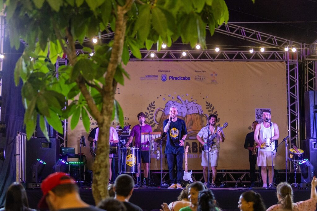 A imagem mostra uma banda tocando em um palco ao ar livre à noite. O palco é decorado com um banner amarelo com imagens de cerveja e texto em português. Há vários músicos no palco, incluindo um vocalista principal, um guitarrista, um baterista e um músico tocando um instrumento de percussão. O público está em primeiro plano, assistindo à apresentação. A iluminação do palco é feita com luzes coloridas e holofotes. Há árvores com folhas verdes em primeiro plano, parcialmente obscurecendo a visão do palco. O evento parece ser um festival ou show em uma área pública.