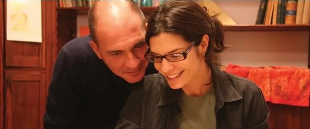 A imagem mostra um close-up de um homem e uma mulher com um fundo desfocado. O homem, à esquerda, está de perfil, com a cabeça inclinada para a direita, olhando para a mulher. Ele tem a cabeça raspada e parece estar sorrindo levemente. Ele está vestindo uma camisa preta de manga comprida. A mulher, à direita, está virada para a frente, mas com o olhar ligeiramente para baixo, como se estivesse olhando para algo que está segurando ou vendo. Ela usa óculos de armação escura e tem um sorriso no rosto. Ela está vestindo uma camisa verde por baixo de uma jaqueta escura. Seus cabelos escuros estão presos em um rabo de cavalo. O fundo sugere que eles estão em um ambiente interno, possivelmente uma sala de estar ou escritório, com estantes de livros visíveis atrás deles. Há também uma peça de tecido laranja vibrante em uma prateleira. A iluminação é suave, focando nos rostos do casal.