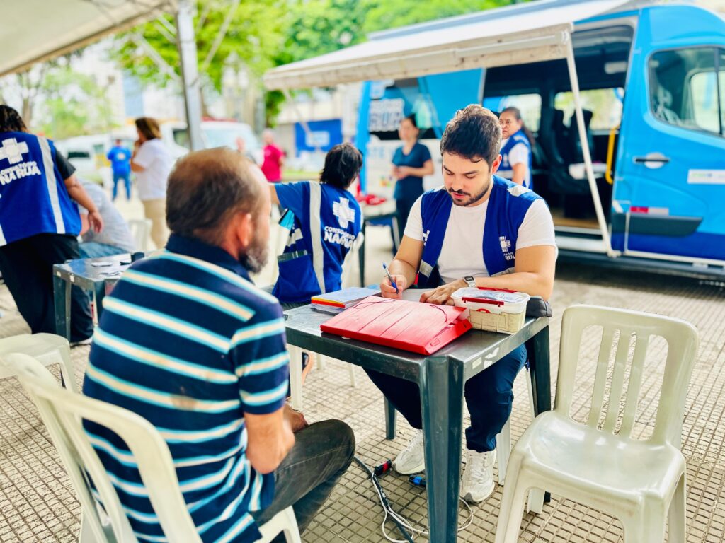 A imagem mostra uma cena ao ar livre, possivelmente um evento de saúde ou serviço público. No centro, um homem com barba está sentado em uma cadeira, de costas para o observador, usando uma camiseta listrada azul e branca. Ele está de frente para uma mesa onde um homem mais jovem, vestindo um colete azul sobre uma camiseta branca e calças azuis, está escrevendo em um papel. O homem mais jovem parece estar preenchendo formulários ou registrando informações. Há uma pasta vermelha e uma caixa pequena sobre a mesa. Ao fundo, uma van azul com o letreiro "RUA" é visível. Várias pessoas estão presentes, algumas usando coletes azuis com um símbolo de cruz branca e a inscrição "CONSULTORIO NA RUA". Uma mulher com um colete semelhante está atrás da van. Outras pessoas estão em segundo plano, algumas em pé e outras sentadas. O ambiente é sombreado por uma cobertura, e o chão é pavimentado com um padrão quadriculado. Há árvores e vegetação ao redor, sugerindo um espaço externo.