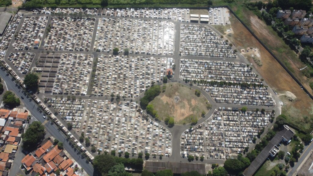 A imagem apresenta uma vista aérea de um extenso cemitério urbano, organizado em seções retangulares com fileiras de túmulos predominantemente brancos ou em tons claros. Caminhos pavimentados cruzam o espaço, facilitando o acesso entre as áreas. No centro, destaca-se uma área circular com uma pequena estrutura — possivelmente uma capela ou monumento — cercada por vegetação, criando um ponto focal sereno.