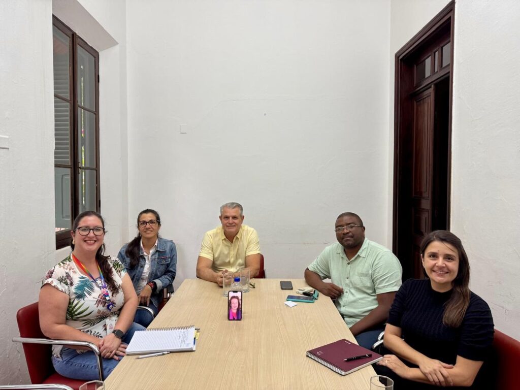 A imagem mostra um grupo de cinco pessoas sentadas ao redor de uma mesa de madeira em uma sala clara. À esquerda, uma mulher com óculos e um colar colorido sorri para a câmera. Ao lado dela, uma mulher com óculos usa uma jaqueta jeans. No centro, um homem de camisa amarela está sentado. À sua direita, um homem de camisa verde clara olha para a câmera. Finalmente, à direita, uma mulher com um vestido preto sorri. Na mesa, há garrafas de água, telefones celulares, cadernos e um pequeno tablet com uma imagem de outra pessoa. A sala tem paredes brancas e uma porta de madeira escura à direita. Há também uma janela com persianas à esquerda.