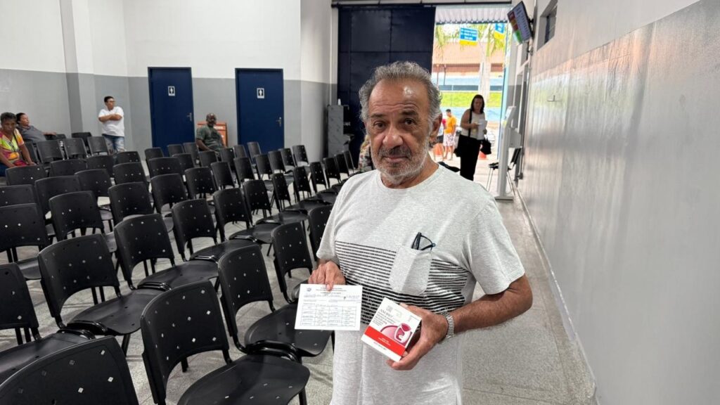 A imagem mostra um homem de meia-idade, com cabelos grisalhos e barba, em pé em um corredor com cadeiras pretas alinhadas. Ele veste uma camiseta cinza com listras pretas no peito e um óculos de sol no bolso. O homem segura um papel e uma pequena caixa vermelha nas mãos. Ao fundo, há portas azuis com símbolos de banheiro, outras pessoas sentadas e em pé, e uma área externa com vegetação. A iluminação é clara, sugerindo um ambiente interno como uma sala de espera ou um centro de atendimento.