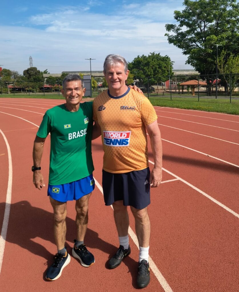 Dois homens estão em uma pista de corrida, com a câmera posicionada em um ângulo que mostra o corpo inteiro deles.O homem à esquerda está vestindo uma camiseta verde com a palavra "BRASIL" em letras brancas e a bandeira brasileira ao lado. Ele também está usando shorts azuis curtos e tênis pretos com detalhes em laranja. Ele tem cabelo grisalho curto e está sorrindo para a câmera.O homem à direita está vestindo uma camiseta laranja com um padrão de escamas e a inscrição "WORLD TENNIS" em letras brancas e azuis. Ele também está usando shorts azuis escuros e tênis pretos com meias brancas. Ele tem cabelo grisalho mais comprido e está sorrindo para a câmera, com o braço esquerdo apoiado no ombro do homem à esquerda.Ambos os homens parecem estar em boa forma física e estão em uma pista de corrida com um céu azul e algumas árvores ao fundo.