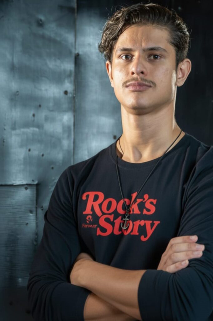 A imagem mostra um close-up de um jovem homem com braços cruzados, vestindo uma camiseta preta com a inscrição "Rock's Story" em vermelho. Ele tem cabelo escuro ondulado, um bigode fino e usa um colar com um pingente de âncora. O fundo é uma parede escura e texturizada.